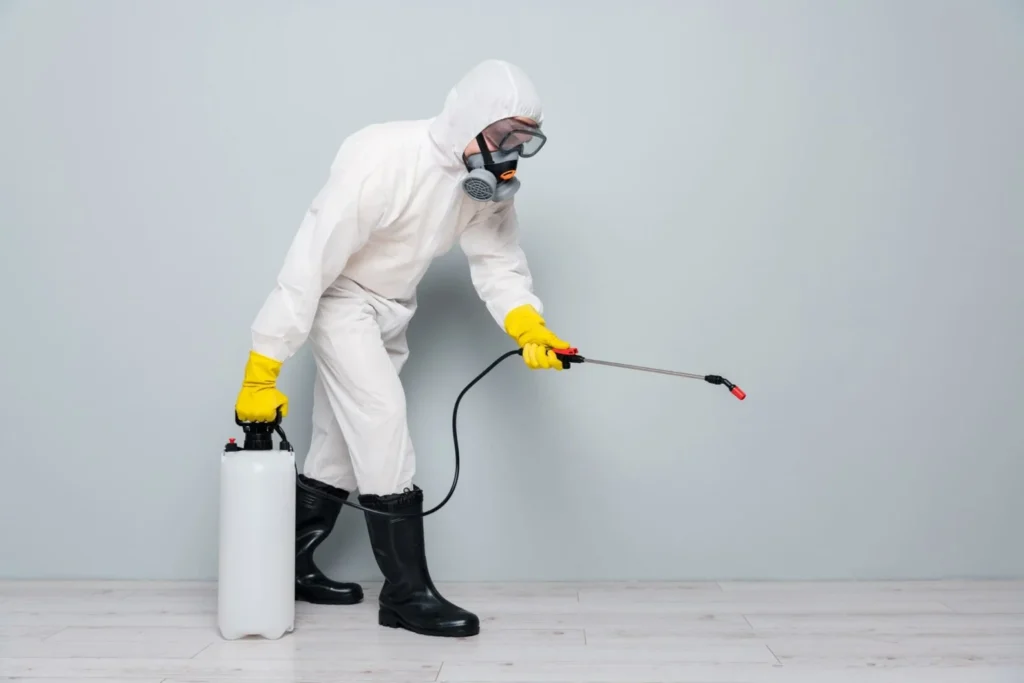 A pest control specialist in a full white hazmat suit, respirator mask, safety goggles, and yellow rubber gloves using a pressurized pump sprayer. The technician is applying a liquid treatment to a floor surface against a plain grey wall background.