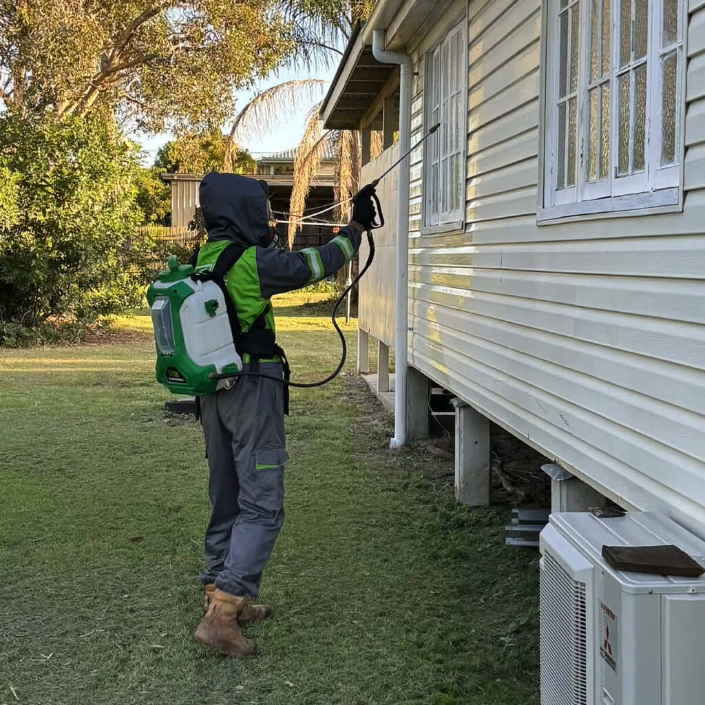 A technician in full protective gear using a backpack sprayer to apply flea treatment to the exterior walls of a home.