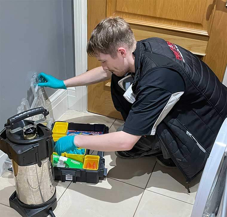 A pest control technician in a black uniform kneeling on a tiled floor while organizing equipment and bait stations from a professional toolkit near an interior doorway.
