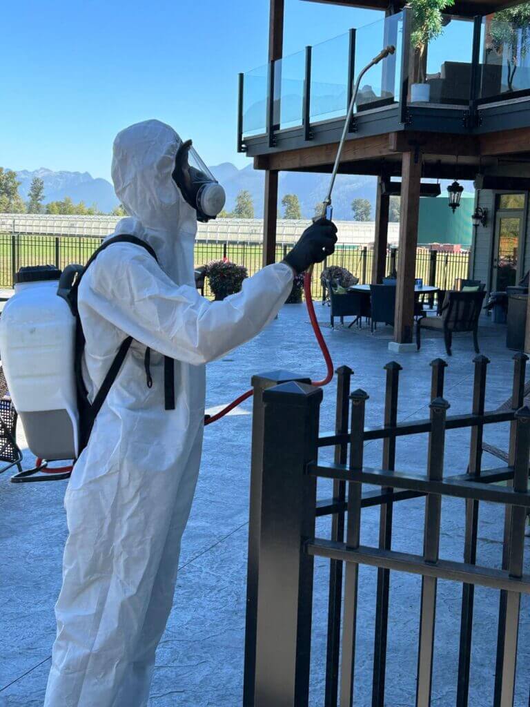 A pest control professional wearing a white protective hazmat suit, respirator mask, and backpack sprayer, treating the exterior of a residential balcony and deck area.